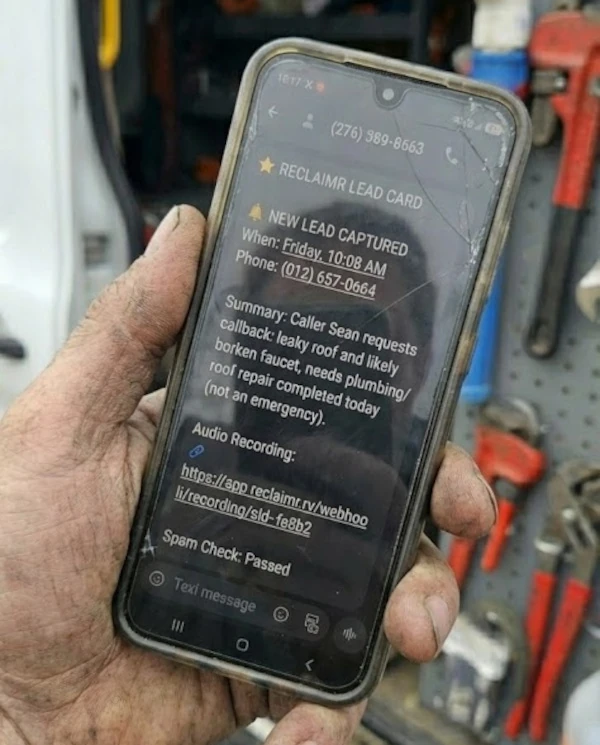 A grease-covered hand holding a phone showing a Reclaimr lead card: new lead captured, the caller's number, a summary of the job, and a link to the call recording.